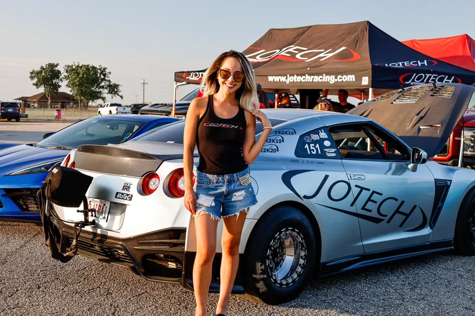 Woman posing with a silver Jotech Racing Nissan GT-R drag car at an outdoor car event with Jotech tents in the background.