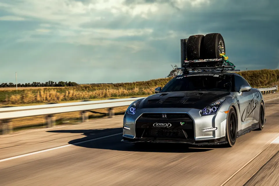 A silver Jotech Nissan GT-R with a carbon hood and roof rack carrying drag tires drives on a highway.
