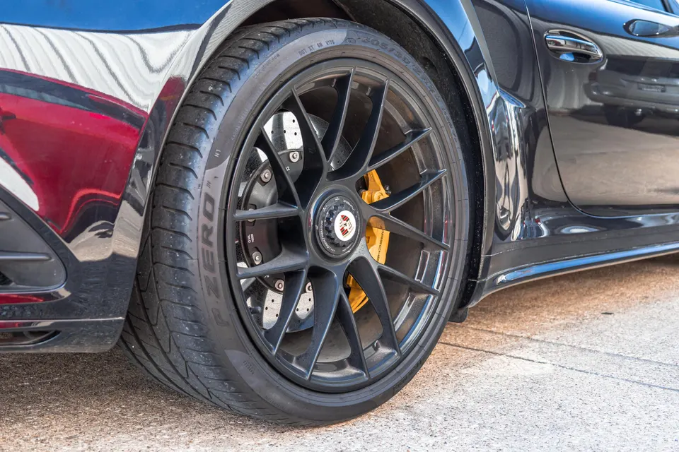 A close-up of a black Porsche 911's wheel, featuring a multi-spoke black wheel, Pirelli P Zero tire, and yellow brake caliper.