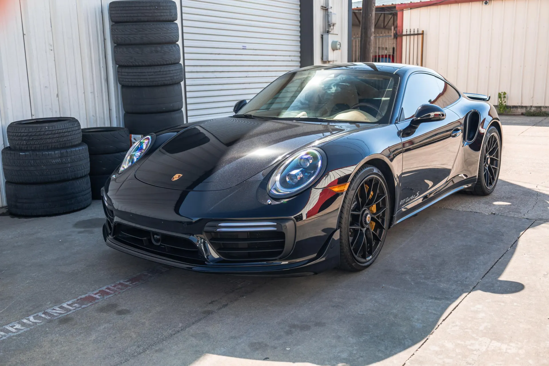 Black Porsche 911 Turbo S parked outside a shop with stacks of tires in the background on a sunny day.