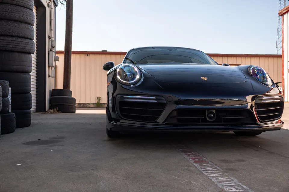 A black Porsche 911 sports car parked outside a shop with stacks of tires in the foreground.
