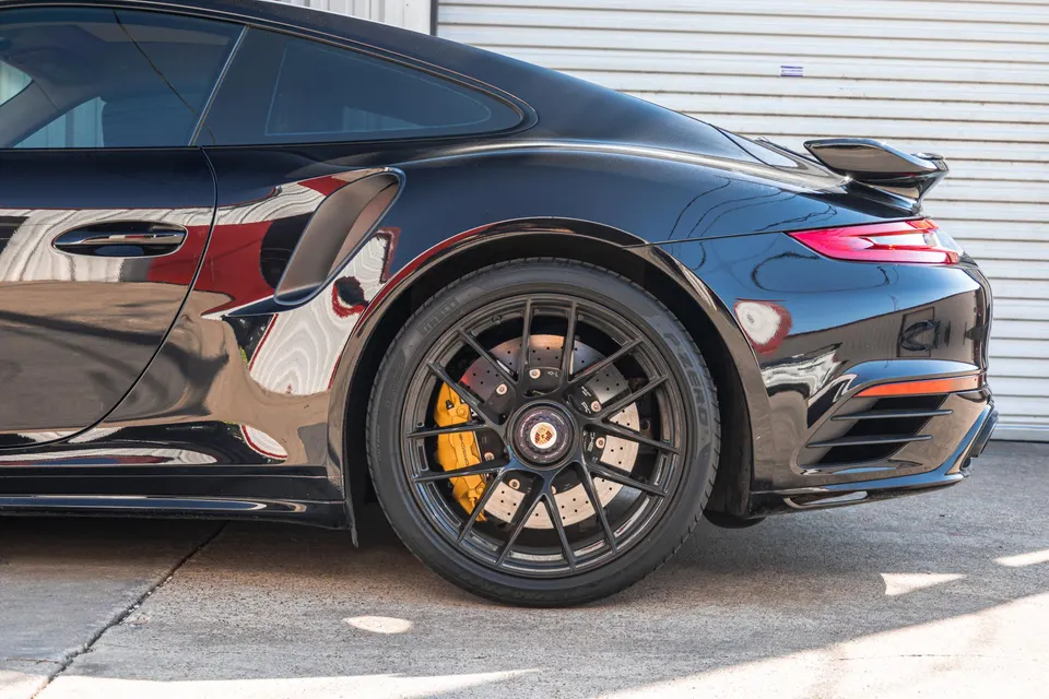 A black Porsche 911 with black multi-spoke wheels and yellow brake calipers parked in front of a garage.