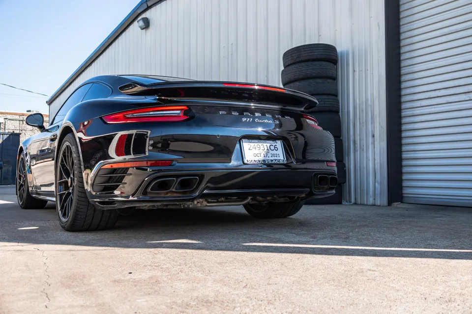 Rear view of a black Porsche 911 Turbo S parked outside a shop, showcasing its exhaust system and rear styling.