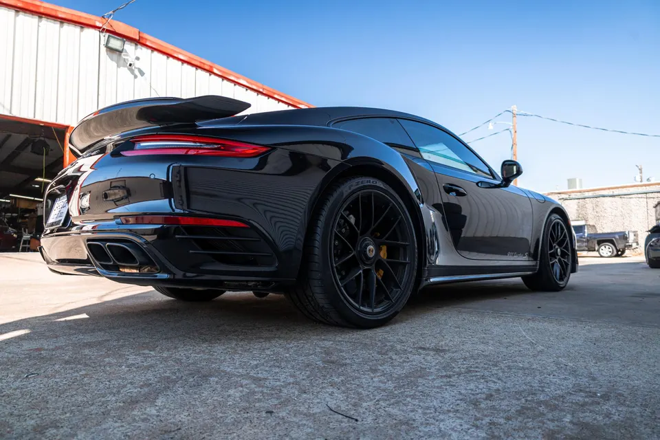 Black Porsche 911 Turbo S parked outside a garage, showcasing its rear and side profile with aftermarket wheels.