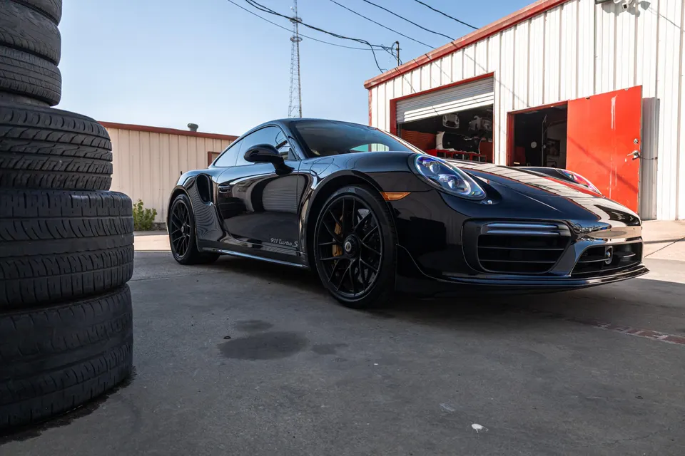 Black Porsche 911 Turbo S parked outside a shop with stacks of tires in the foreground, under a clear sky.