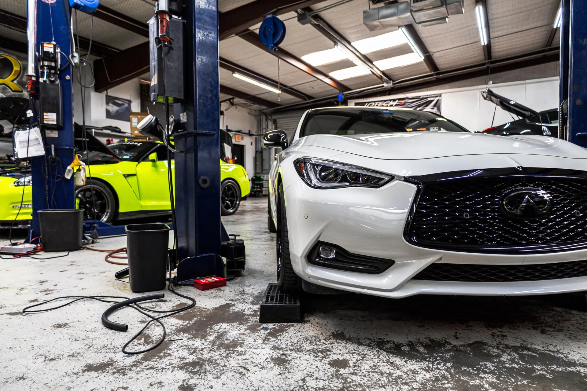 A white Infiniti Q60 and a neon green Nissan GT-R inside a performance tuning shop with car lifts.