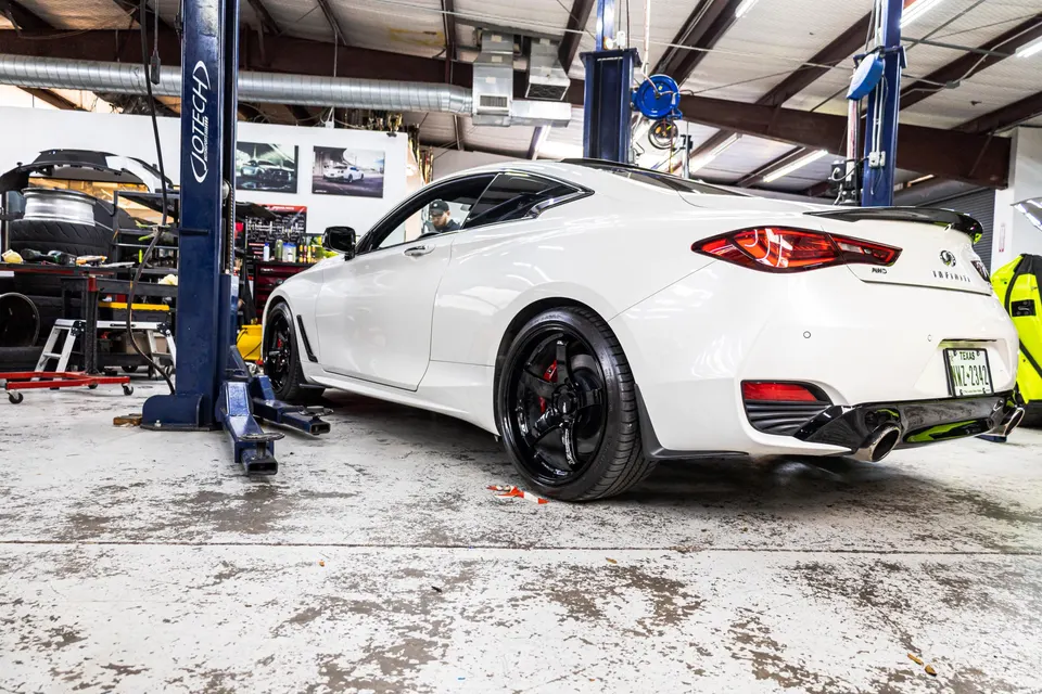 White Infiniti Q60 on a lift in a performance tuning shop, showcasing aftermarket wheels and quad exhaust tips.