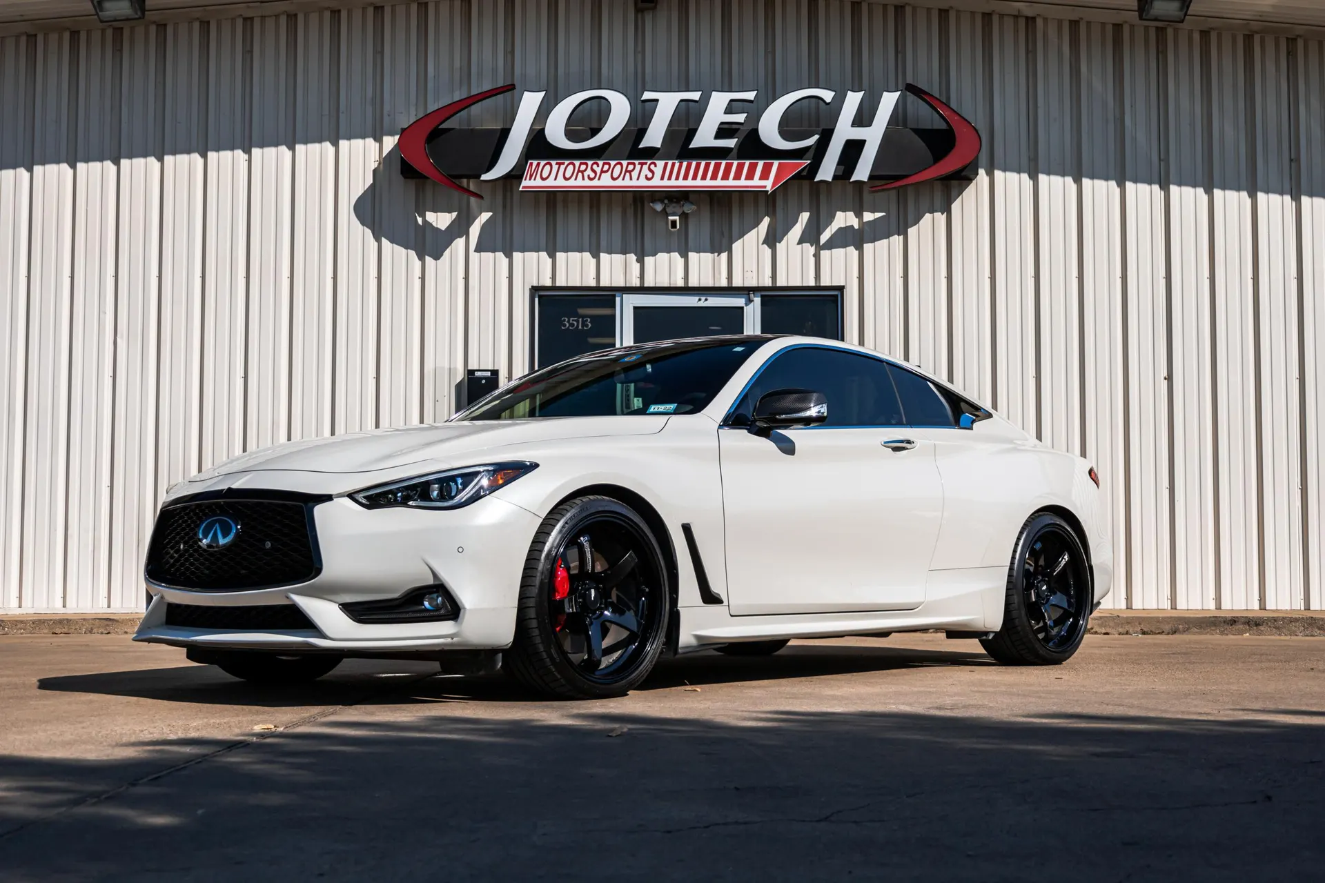 White Infiniti Q60 coupe with black aftermarket wheels and red brake calipers parked in front of Jotech Motorsports.