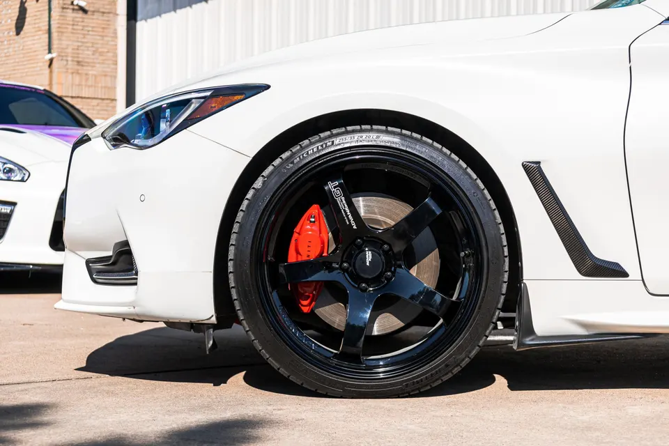 A close-up of a white Infiniti Q60's front wheel, featuring black Advan GT wheels, red Infiniti calipers, and carbon fiber fender trim.