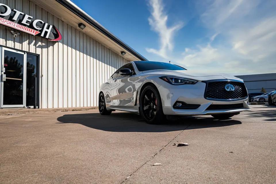 A white Infiniti Q60 coupe with black wheels parked outside the Jotech Motorsports building on a sunny day.