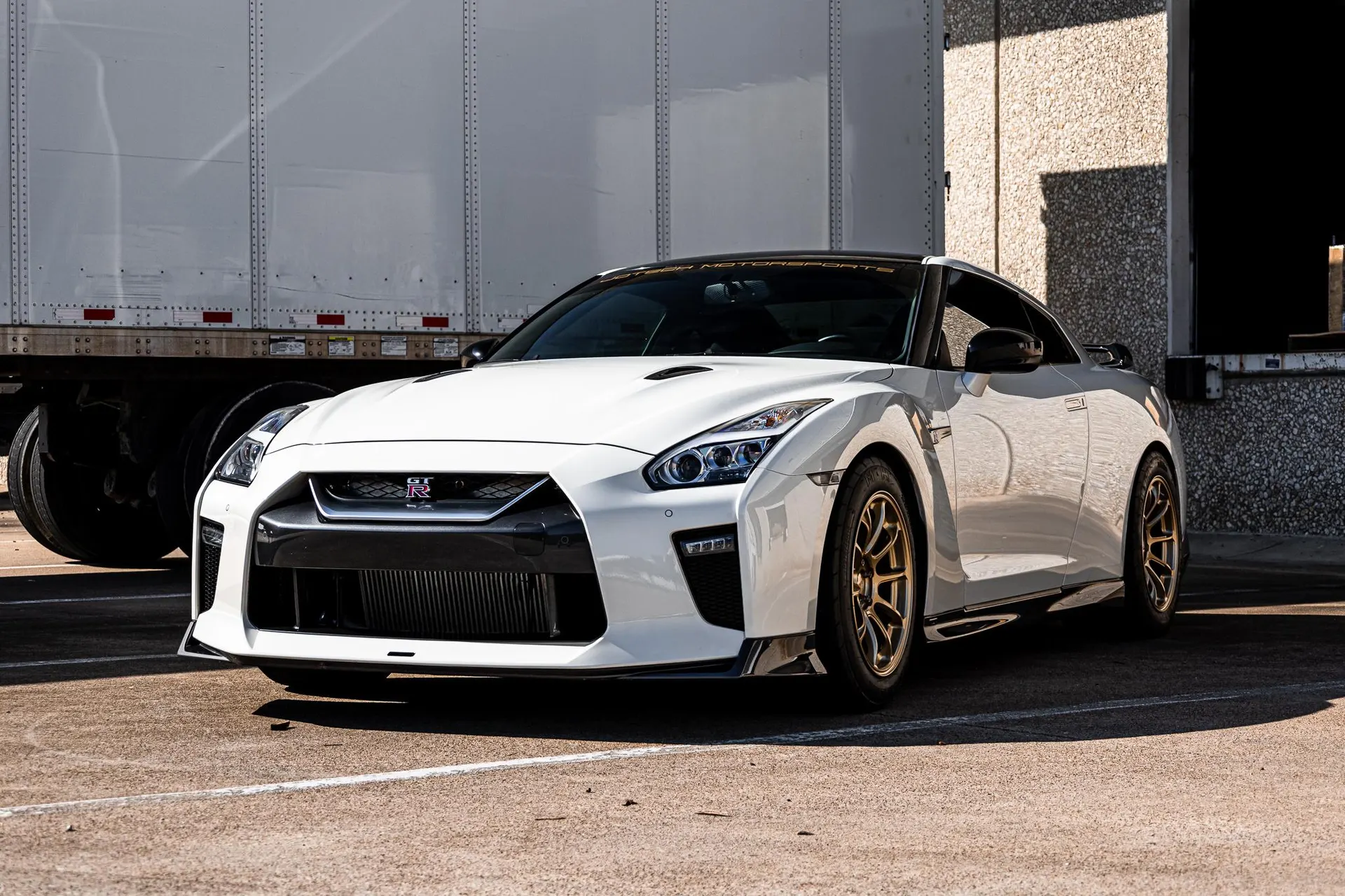 A white Nissan GT-R R35 with bronze aftermarket wheels parked in front of a white truck trailer and building.