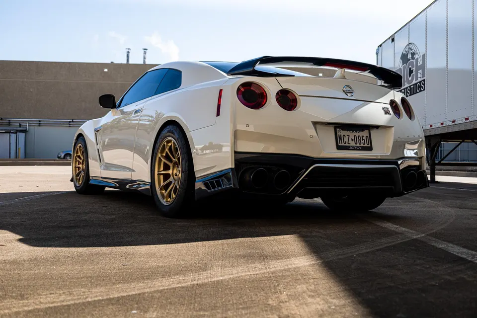 White Nissan GT-R with gold aftermarket wheels, carbon fiber aero, and quad exhaust tips parked in a lot.