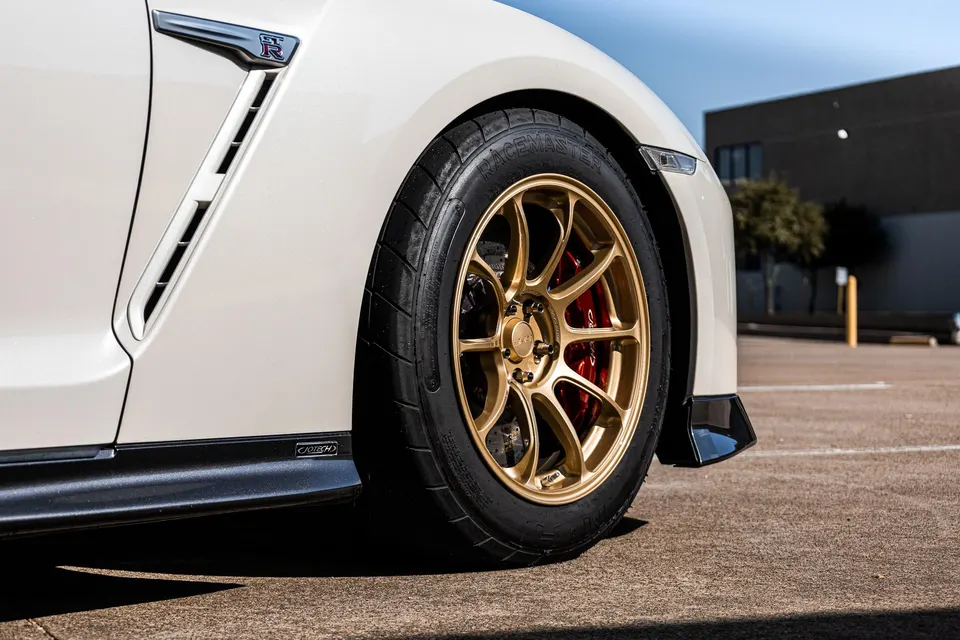 A close-up of a white Nissan GT-R's front wheel, featuring gold multi-spoke wheels, red brake calipers, and Racemaster tires.