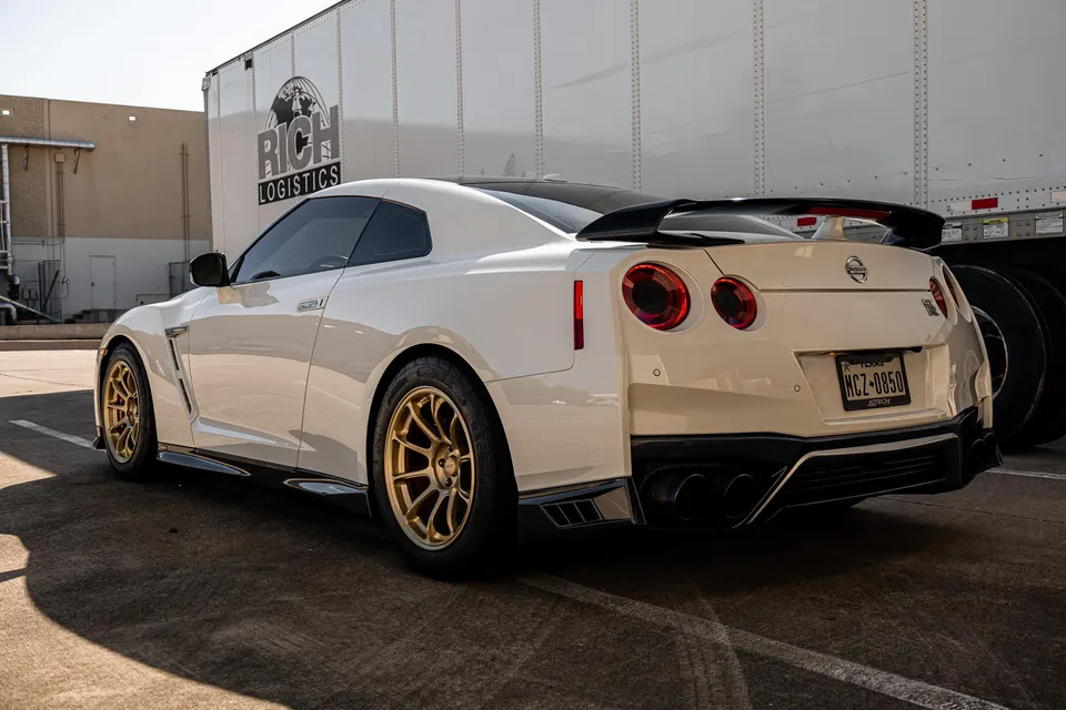 A white Nissan GT-R with gold aftermarket wheels and a black rear spoiler parked in a lot next to a white truck.