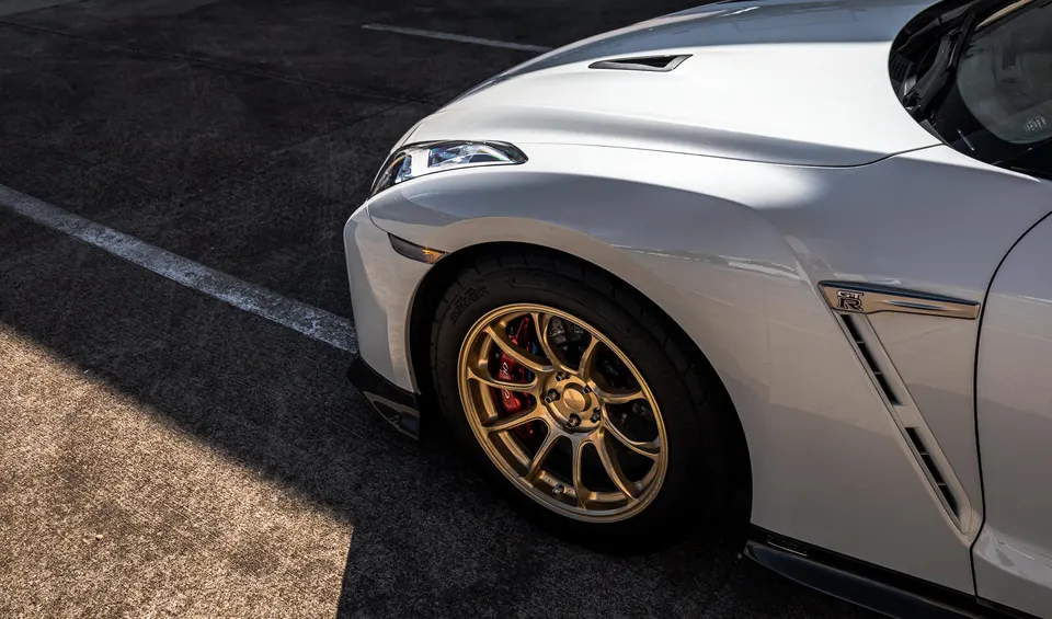 Close-up of a white Nissan GT-R's front fender, showcasing gold aftermarket wheels and red brake calipers.