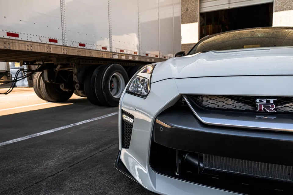 A white Nissan GT-R with a visible intercooler parked next to a semi-trailer in an industrial setting.
