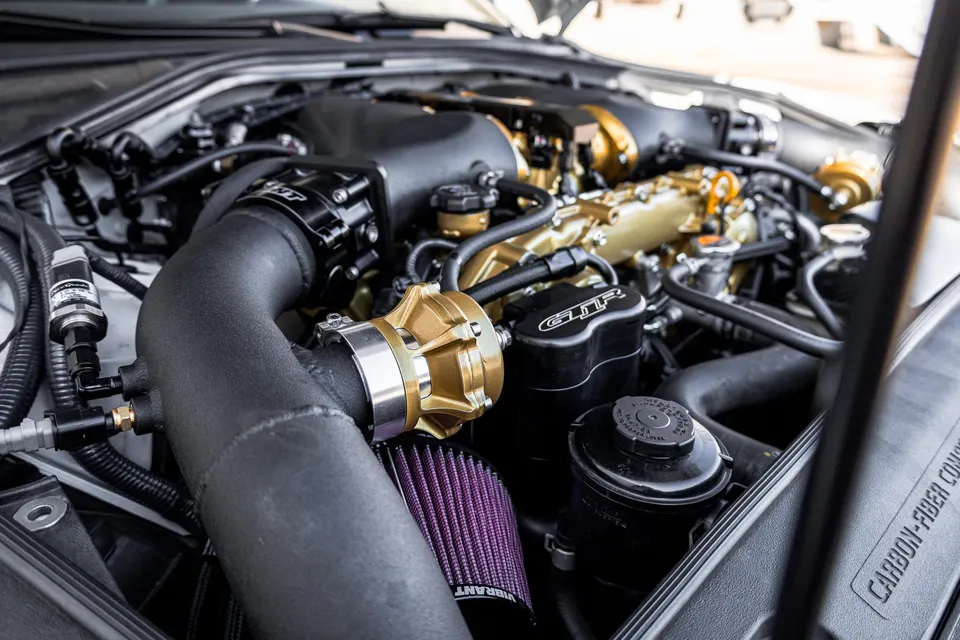 Close-up view of a modified Nissan GT-R engine bay featuring a gold blow-off valve, aftermarket intake, and gold engine covers.