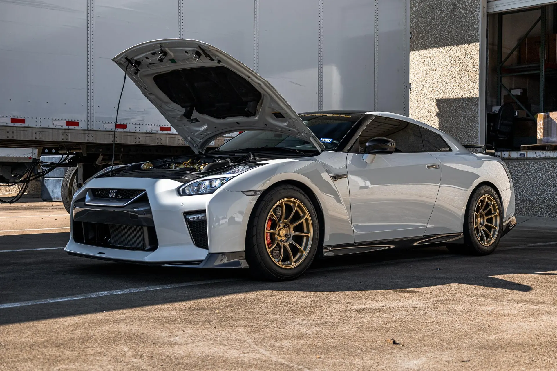 A white Nissan GT-R with its hood open, parked outside a building with a semi-trailer in the background.