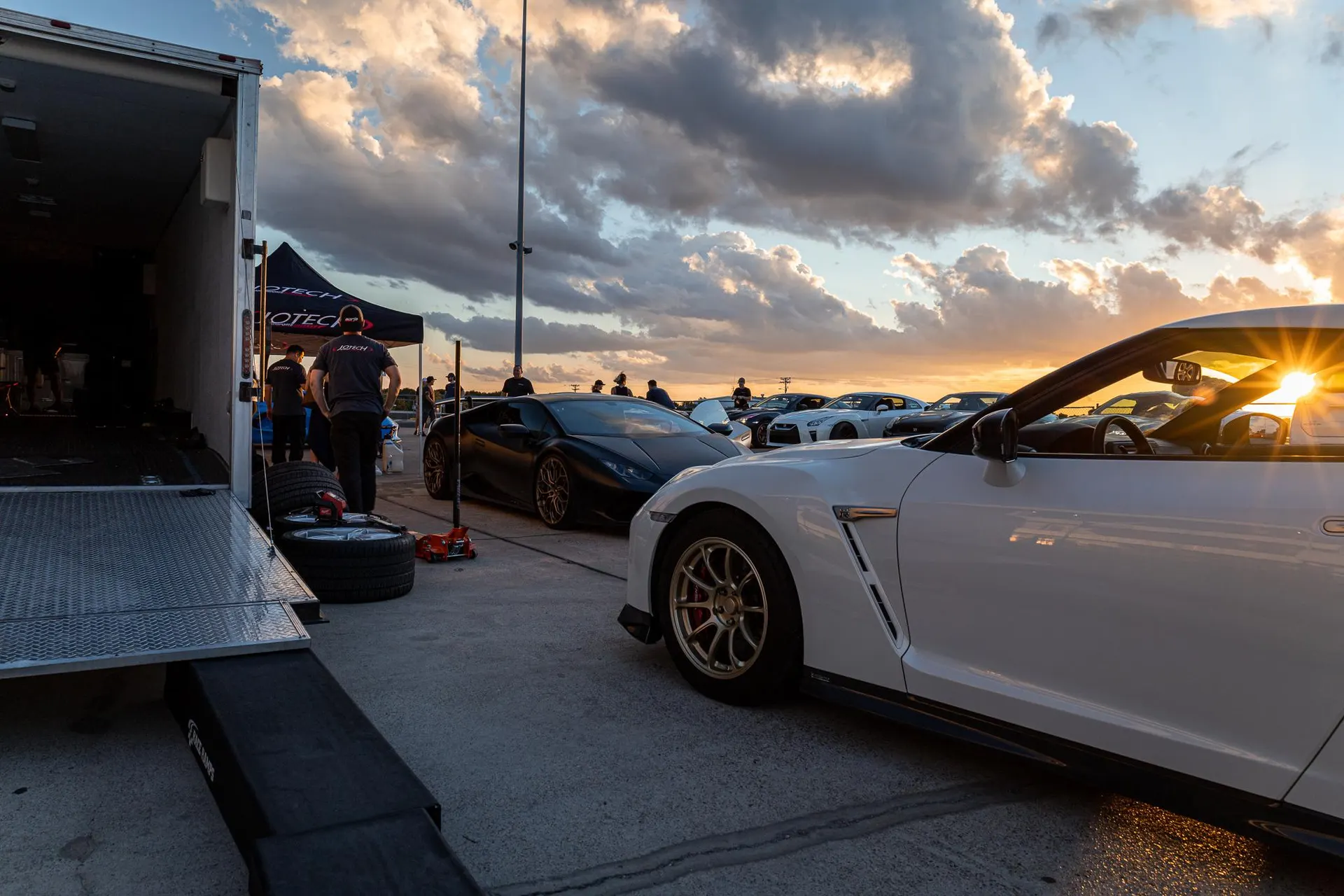 Nissan GT-R and Lamborghini Huracan parked at a track event during sunset with a trailer and people in the background.