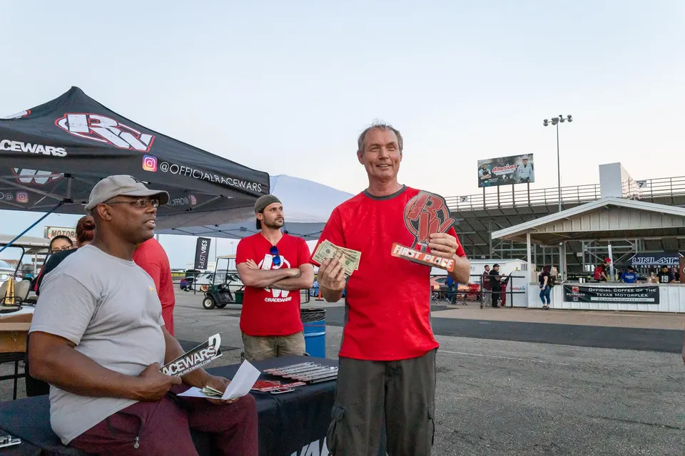 People celebrating at a Racewars event, with one man holding a 'First Place' trophy and cash prize at a drag strip.