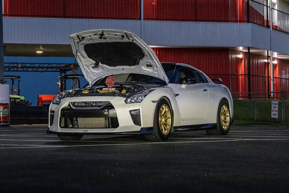 White Nissan GT-R with hood open, revealing a custom engine bay and a first place trophy, parked at a track event.