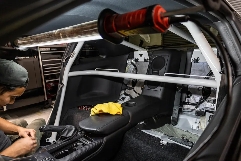 Mechanic working on the interior of a car with a white roll cage installed and rear seats removed in a performance shop.