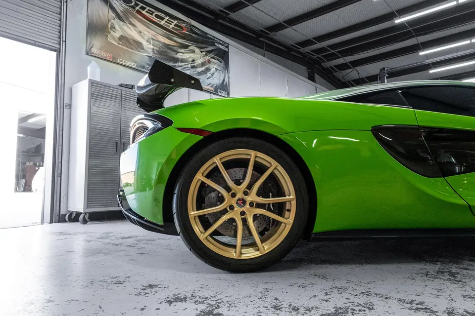 Green McLaren supercar with gold aftermarket wheels and a carbon fiber rear wing parked inside a performance tuning shop.