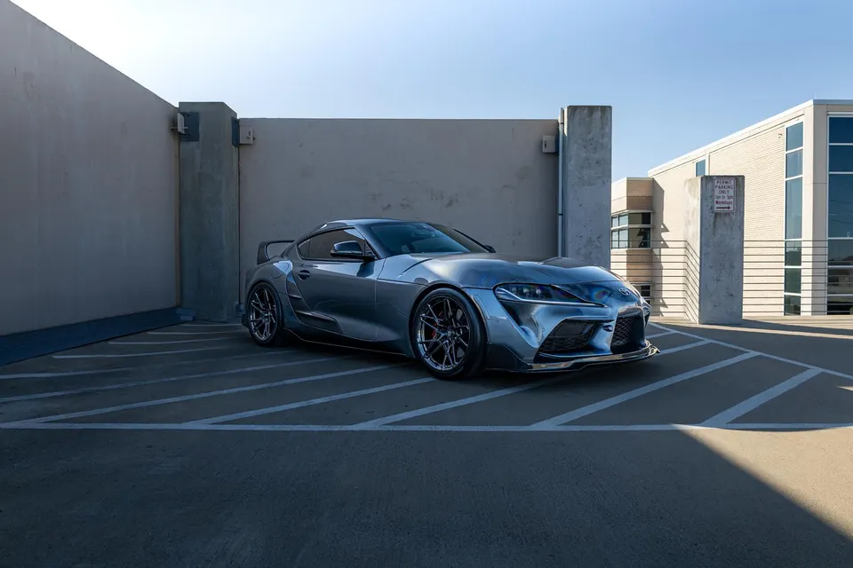 A modified grey Toyota Supra with a widebody kit and aftermarket wheels parked in a modern concrete setting.