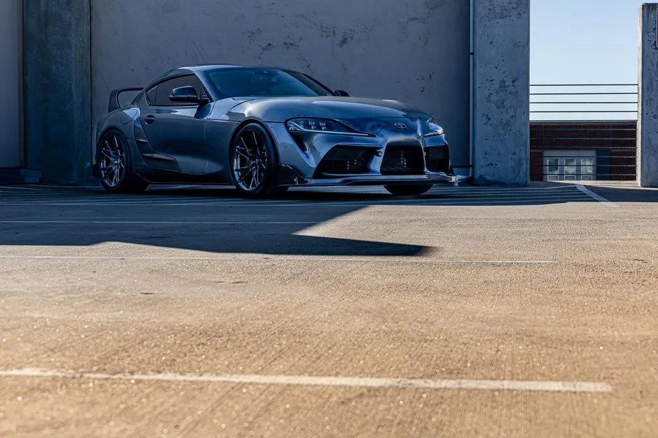 Grey widebody Toyota Supra with aftermarket wheels, front splitter, and rear wing parked in a concrete structure.