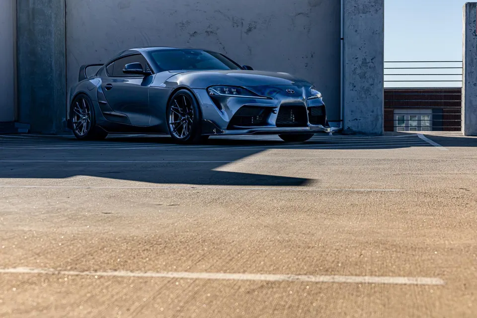 A grey widebody Toyota Supra MK5 with aftermarket wheels and a rear wing parked in a concrete parking structure.