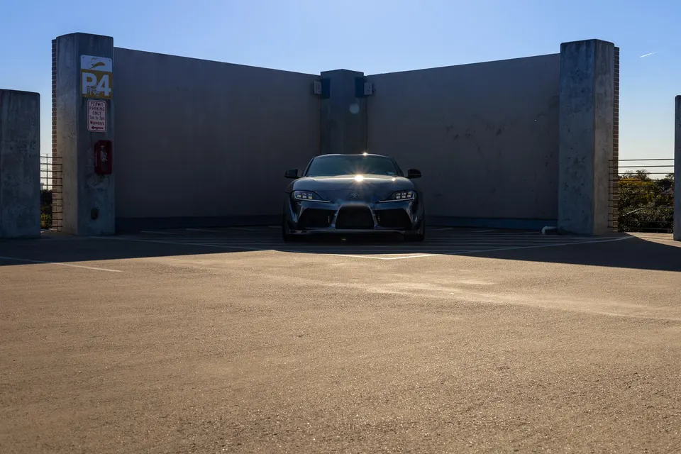 Front view of a grey Toyota Supra parked in a concrete parking structure under a clear blue sky.