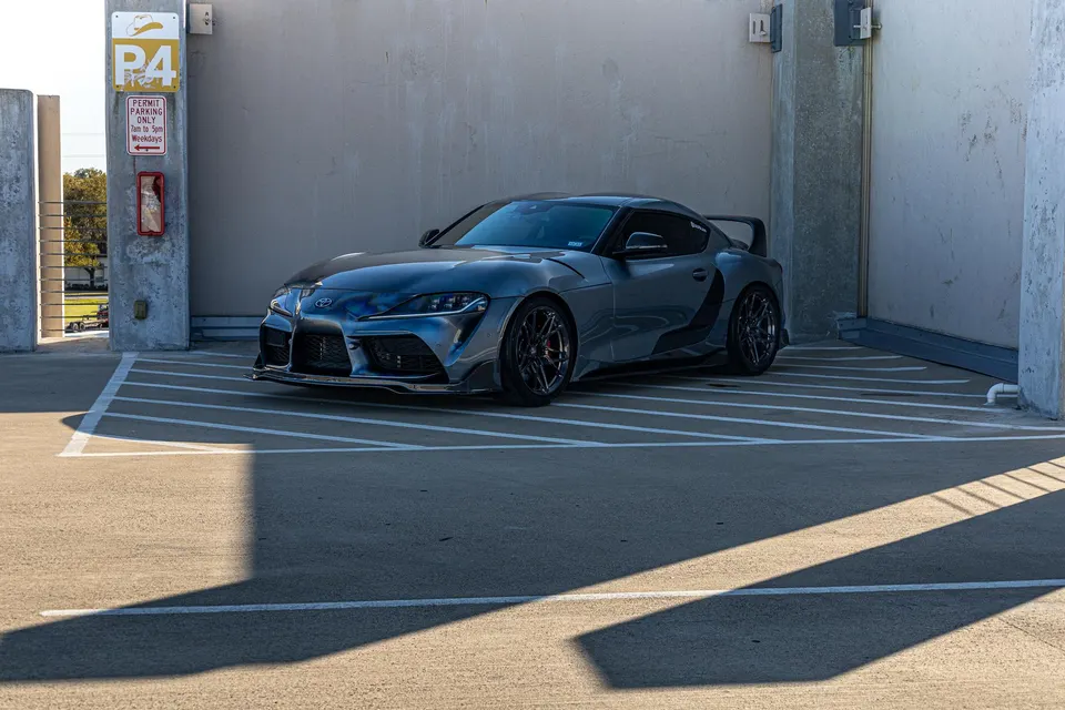 A grey Toyota Supra MK5 with aftermarket wheels and a rear wing parked in a concrete parking garage.