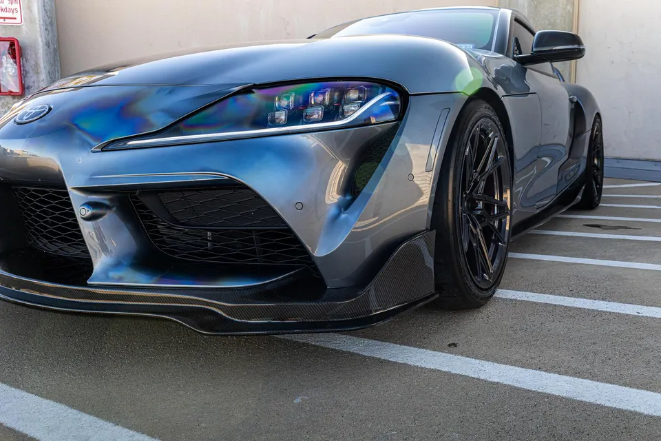 Custom grey Toyota Supra with iridescent reflections, featuring a carbon fiber front splitter and aftermarket black wheels.