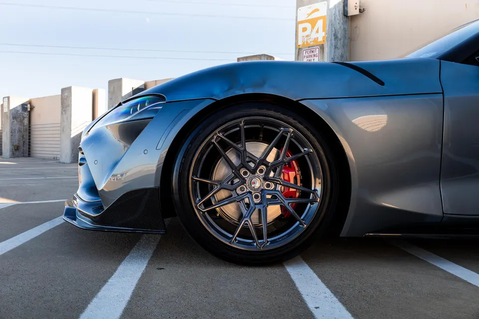 Close-up of a grey Toyota Supra's custom multi-spoke wheel with red brake calipers and carbon fiber front splitter.