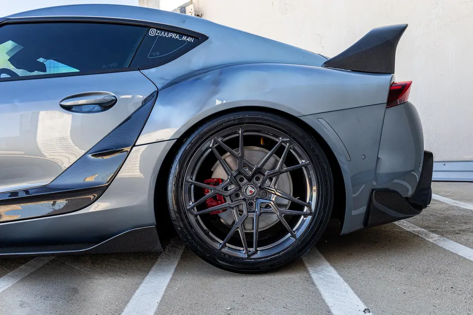 A grey MK5 Supra with custom dark multi-spoke wheels, red brake calipers, and carbon fiber aerodynamic parts, parked outdoors.