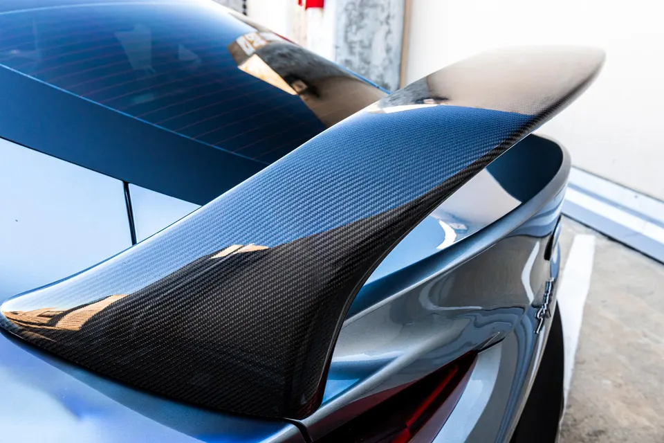 Close-up of a carbon fiber rear spoiler on a blue performance car, showcasing the weave pattern and glossy finish.