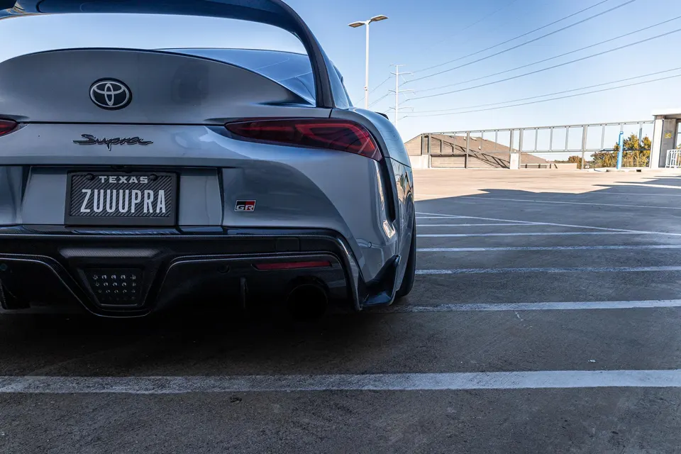 Rear view of a silver Toyota Supra A90 with a custom Texas license plate, showcasing its aggressive rear diffuser and exhaust.