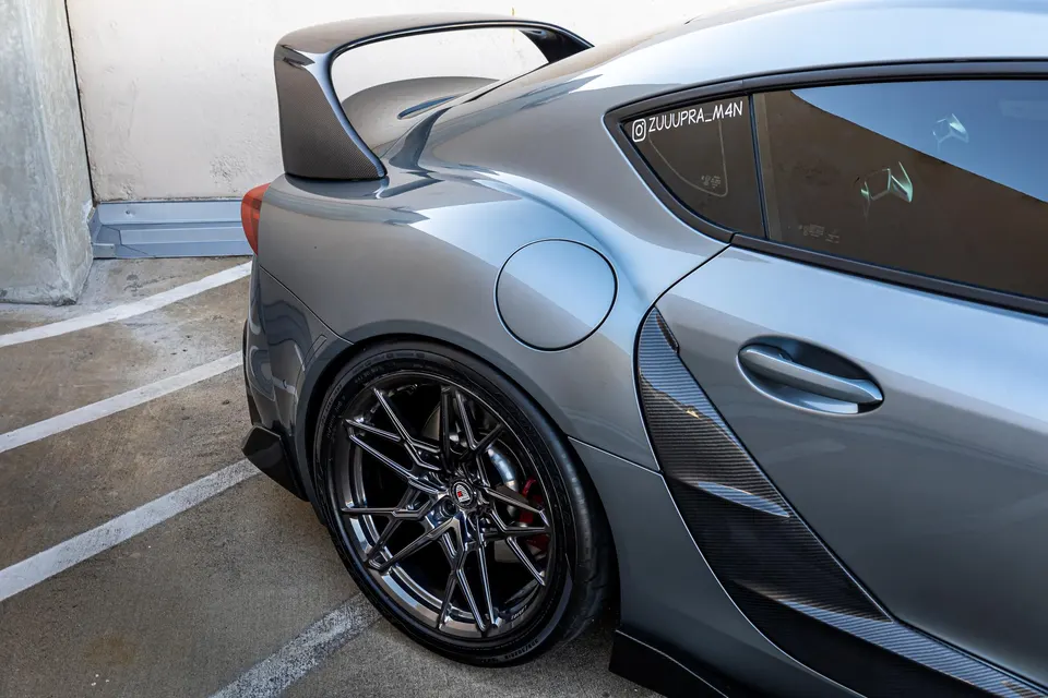 A grey Toyota Supra with aftermarket wheels, a carbon fiber wing, and carbon fiber side vents parked outdoors.