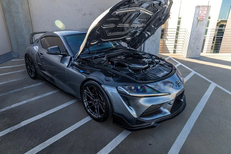 Grey Toyota Supra with hood open showcasing the engine bay and aftermarket wheels parked outdoors.
