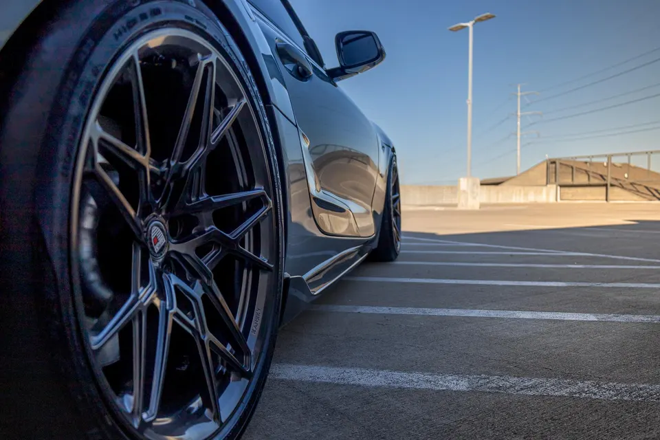 Close-up of a performance car's custom multi-spoke wheel and glossy dark paint, parked on a sunny rooftop.