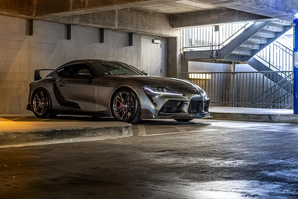 A grey Toyota Supra with aftermarket wheels, a front splitter, and a large rear wing parked in a concrete parking garage.