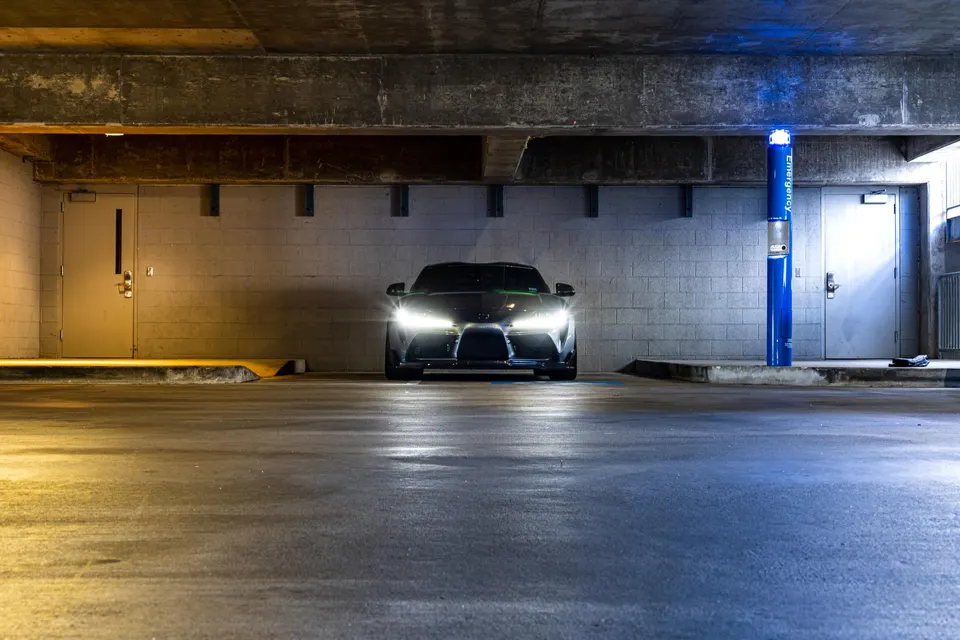 A dark grey Toyota Supra A90 with its headlights on, parked head-on in a concrete parking garage.