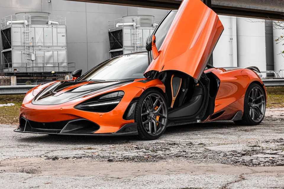 Orange McLaren 720S with butterfly doors open, featuring aftermarket wheels and carbon fiber accents, parked in an industrial setting.