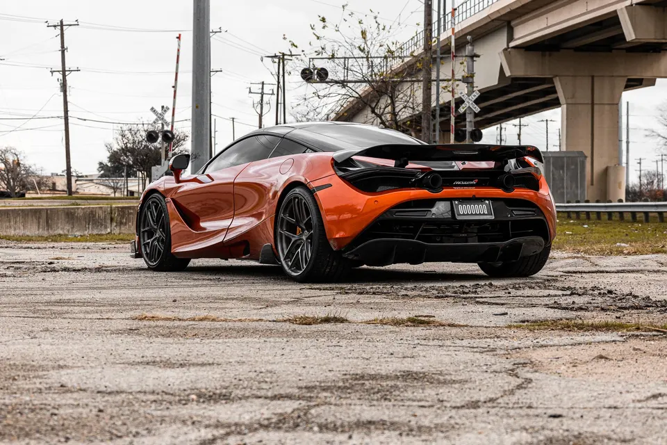 An orange McLaren 720S with aftermarket wheels parked near a railroad crossing and overpass.