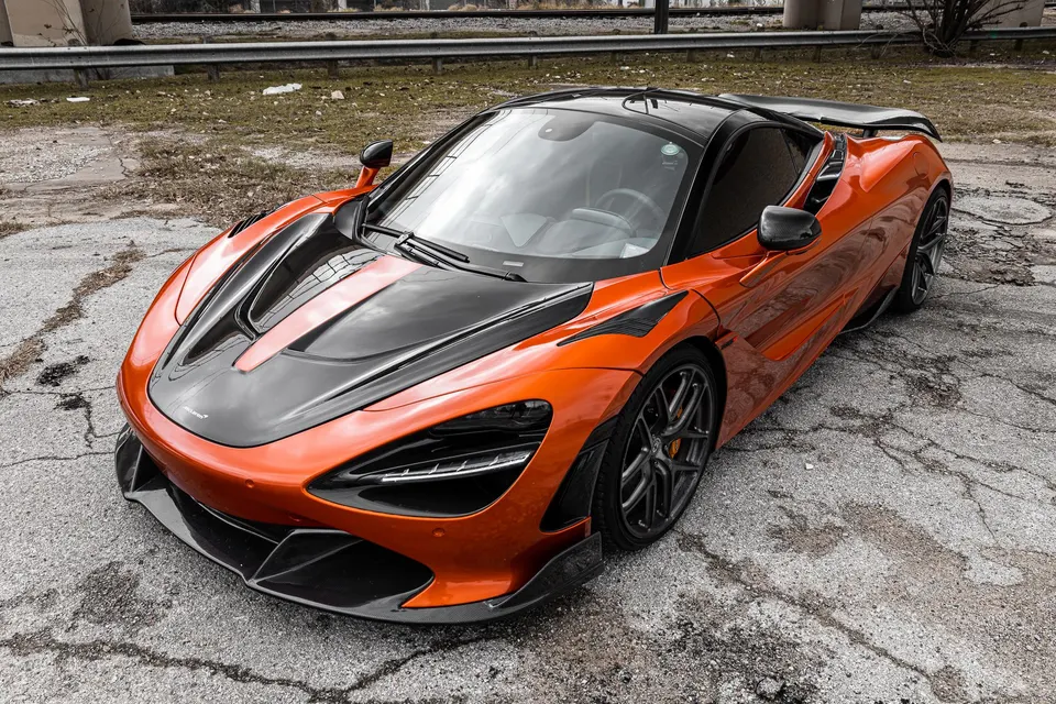 Orange McLaren 720S supercar with black carbon fiber hood and aero parked on cracked asphalt.