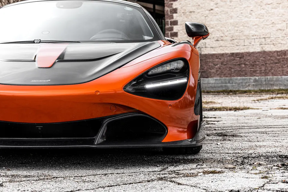 Front view of an orange McLaren 720S with a black carbon fiber hood and front splitter parked on asphalt.