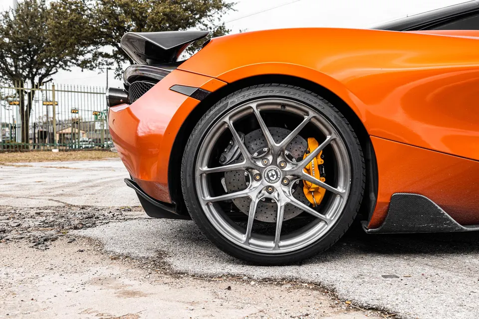 Close-up of an orange McLaren's rear wheel, showcasing HRE wheels, yellow brake calipers, and carbon fiber aero.