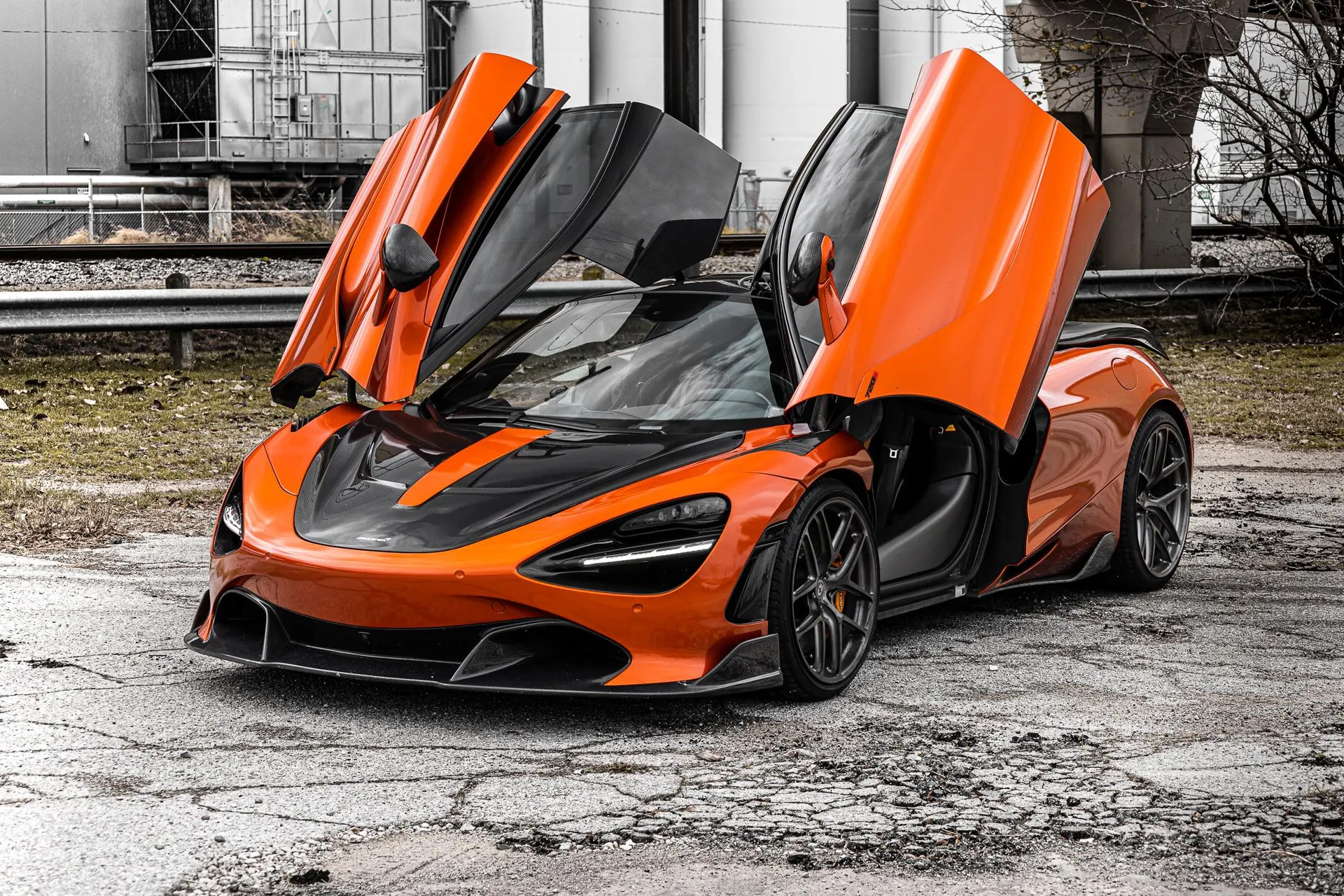 Orange McLaren supercar with butterfly doors open, parked in an industrial setting with buildings in the background.