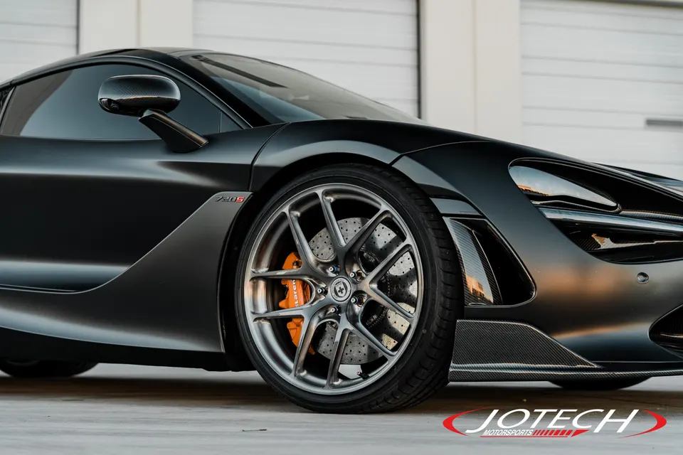 Close-up of a matte grey McLaren 720S showing custom multi-spoke wheels, orange brake calipers, and carbon fiber accents.