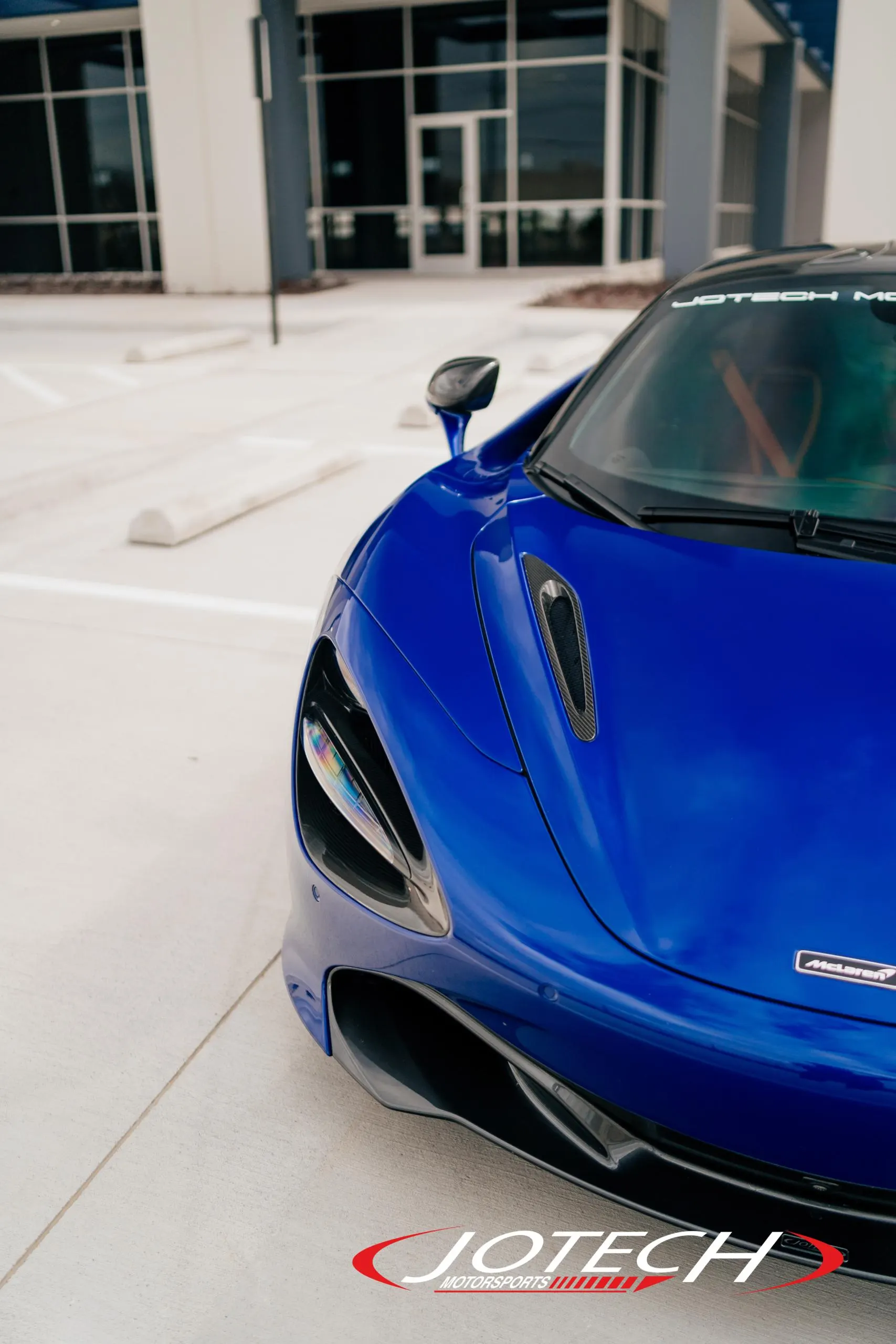 A blue McLaren supercar's front end, showcasing its sleek design, headlight, and carbon fiber accents outside Jotech Motorsports.
